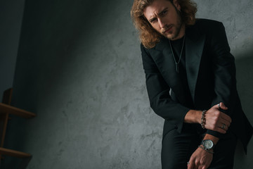 low angle view of tense fashionable businessman in black suit posing in dark near grey wall