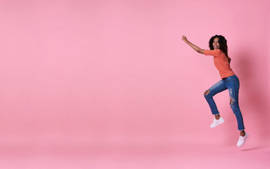 Joyful young african woman in orange shirt jumping and celebrating over pink banner background with copy space.