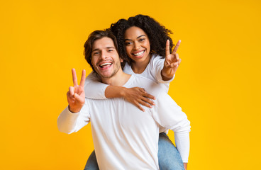 Joyful interracial couple embracing and showing peace sign over yellow background