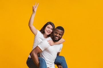 Fototapeta premium Cheerful woman riding her black boyfriend's back and showing peace gesture