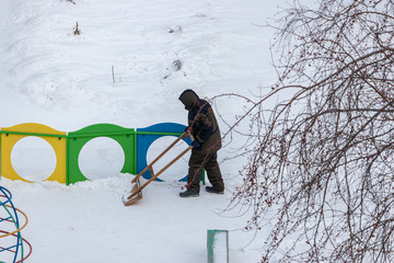 Cleaner cleans snow with a large wooden shovel in the playground