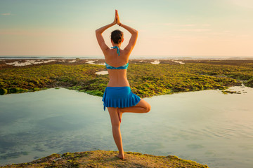 Vrikshasana asana. Young woman practicing tree pose at the beach during sunset. Arms raising with...