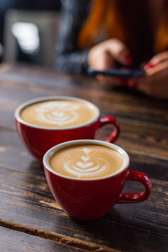 Fototapeta Dwa czerwonej filiżanki kawy na drewnianym tle z latte sztuką. Stolik w kawiarni. Koncepcja łatwego śniadania. Małe i duże ceramiczne kubki. Widok z góry. Dziewczyna z telefonem w tle