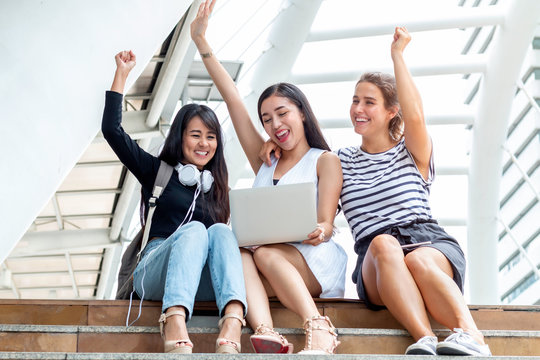 Cheerful multi ethnic friends using laptop computer and sitting on the staircase at university campus.