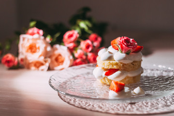 Sponge cake with white cream and roses on top on a transparent plate with a reflection on the table