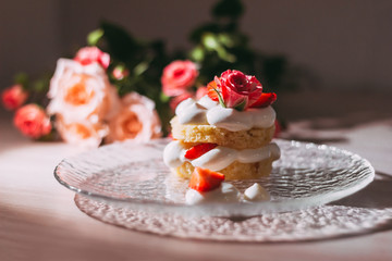 Sponge cake with white cream and roses on top on a transparent plate with a reflection on the table on a background of flowers