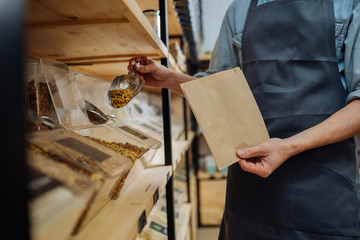 Mixed race male worker shop assistant filling paper bag with oat granola in bulk products in...