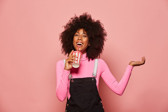Joyful young African American female with curly hair wearing pink turtleneck and black overall holding white and pink striped disposable cup with straw and looking at camera