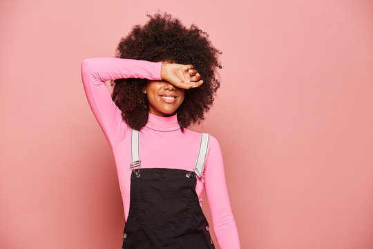 Excited millennial African American female with curly hair wearing pink turtleneck and black overall covering eyes with arm and waiting for surprise while standing against pink background