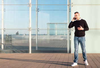 Fototapeta na wymiar Handsome silver fox man talking on mobile phone with glass office building in background 