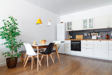 Modern interior of cozy kitchen, dining room, white furniture, wall, black accessories, natural materials, wooden chairs, green flower in pot. Concept scandinavian design apartment of young family