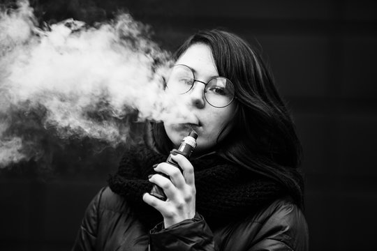 Vape teenager. Young pretty white caucasian girl in a jacket and glasses smoking an electronic cigarette opposite modern brown background on the street in the winter. Deadly bad habit. Black and white