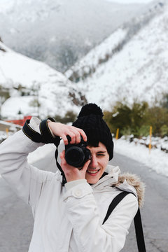 Focused female in white winter jacket with hood and black pants taking picture with camera while standing in middle of asphalt road between snowy mountains