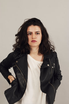 Thoughtful attractive dark haired woman in black biker jacket and looking at camera on gray background in studio