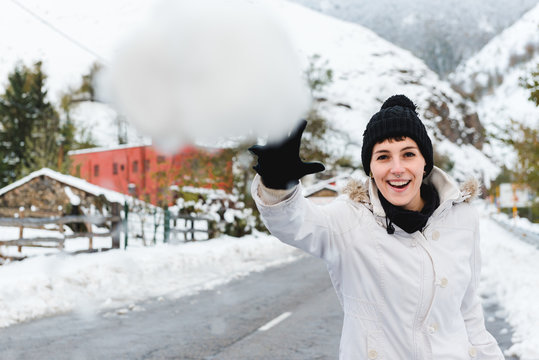 Cheerful female in black knitted hat with pompon and white winter jacket throwing snowball at camera while walking in mountain village
