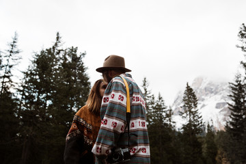 Side view of young couple tourists in stylish casual wear standing and hugging on empty road among pine forest with Dolomites mountains on background at Italy