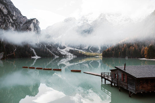 Wooden house on stilts and boats on nebulous lake with reflection of powerful Dolomites mountains at Italy