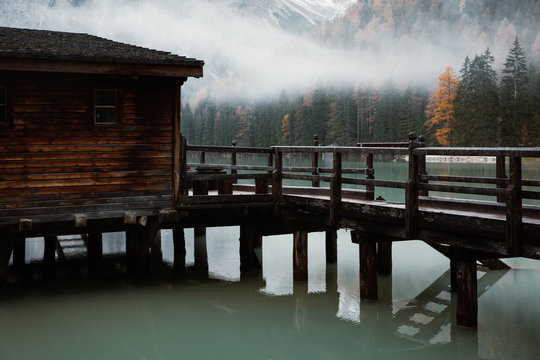 Wooden house on stilts and boats on nebulous lake with reflection of powerful Dolomites mountains at Italy