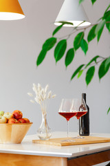 Dark bottle and two glasses of red wine, wooden bowl with apples, decor vase on table in modern kitchen, lamps over table, green leaves. Vertical. Copyspace. Place for text, interior