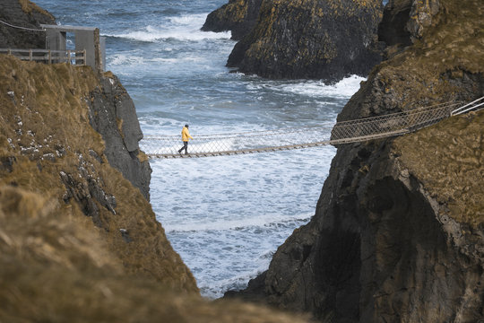 Fototapeta From above side view of traveler passing over Carric a Rede rope bridge suspended between rocky cliffs and sea waves crashing on rocks in background
