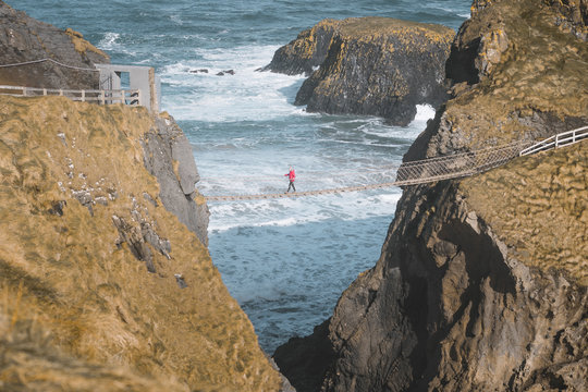 From above side view of traveler passing over Carric a Rede rope bridge suspended between rocky cliffs and sea waves crashing on rocks in background