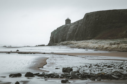 Scenic landscape with Mussenden Temple located on stone cliff at Northern Ireland coastline and stormy sea waves dashing against rocks with grey cloudy sky in background