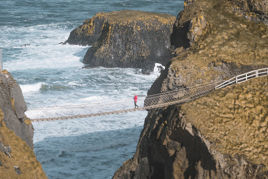 From above side view of traveler passing over Carric a Rede rope bridge suspended between rocky cliffs and sea waves crashing on rocks in background