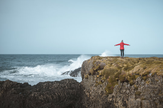 Back view of faceless free female in warm outfit on cliff top enjoying stormy raging surf with spread arms in Ballintoy