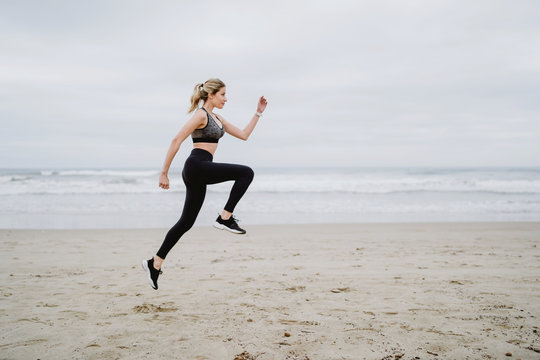 Side view of determined female jogger in stylish sportswear and sneakers doing cardio in jump at empty seashore on cloudy weather