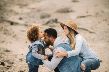 Cheerful multiracial parents in stylish casual clothes holding smiling adorable curly ethnic toddler and having fun at sandy landscape