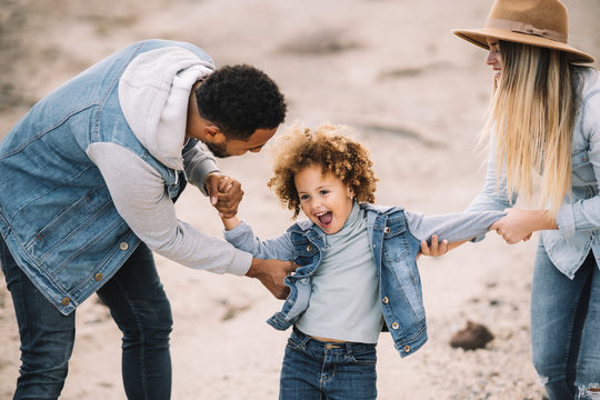Cheerful multiracial parents in stylish casual clothes holding smiling adorable curly ethnic toddler and having fun at sandy landscape