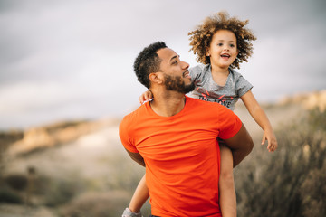 Smiling African American bearded man dressed in sportive clothes carrying cheerful curly active toddler while walking on nature