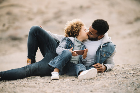 African American bearded casual man lying on sandy ground leaning on elbow and bending leg at knee looking at curly ethnic toddler in denim clothes sitting beside with mobile phone - Powered by Adobe