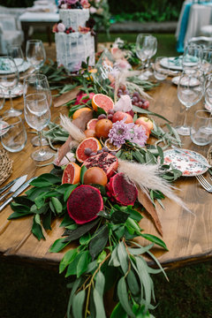 Beautiful garland made from ripe cut tropical fruits and green branches with flowers and decorative plants setting on round wooden wedding table served with plates and glasses 