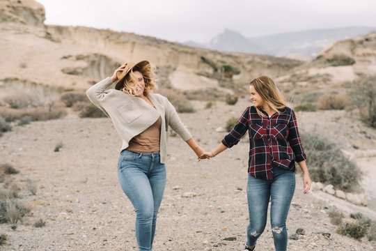 Woman in hat wearing casual clothes tenderly holding hands of female with long straight hair dressed in check shirt smiling on nature