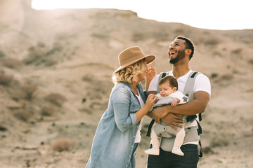 Happy black bearded man carrying little baby and holding hand of blond wife wearing denim shirt jeans and hat while standing on sandy land