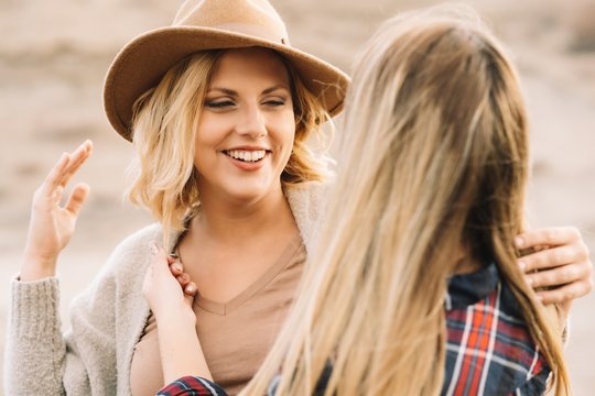 Woman in hat wearing casual clothes tenderly hugging female with long straight hair dressed in check shirt smiling on nature