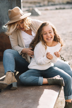 Content woman with blond hair in hat cuddling and tickling daughter in casual clothes while having rest and sitting in old truck body