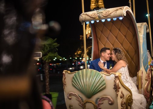 Happily married young man in blue suit and woman in wedding dress looking at each other while taking amusement rides in an embrace with Eiffel Tower on background