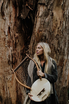 Dreamy charming tender woman with blond hair holding wooden antique musical instrument while standing with closed eyes at old tree trunk