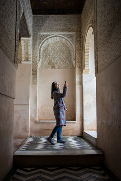Full body side view of woman tourist admiring ornament over arched window inside ancient Islamic palace taking picture with small camera device