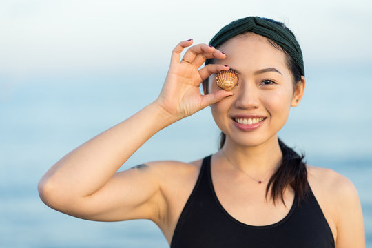 Cheerful smiling young Asian female in sport shirt looking at camera and covering eye with sea shell while standing on beach with blurred blue water and sky in background