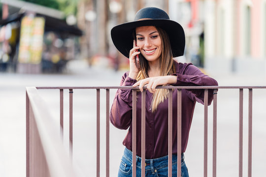 Content long haired lady in fashionable black hat and shirt leaning on fence while calling on mobile phone and looking away at city street