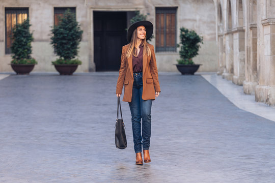 Excited female in stylish casual wear and black hat walking and looking away on sidewalk among old buildings