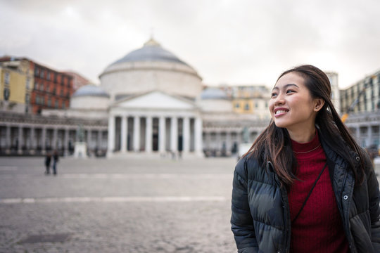 Pleased Asian female traveler in warm clothing smiling with historical beautiful Basilica of San Francisco de Paula on blurred background at Plebiscite Square at Naples