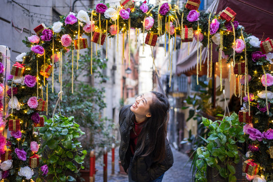 Overjoyed Asian female tourist in casual clothing standing under festive arch with flowers and fairy lights and laughing at Spanish Quarter at Naples