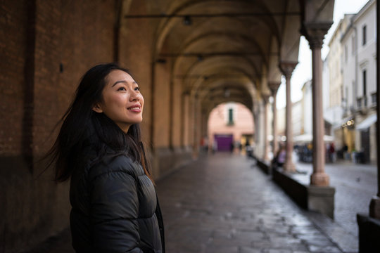Content Asian resting female smiling wile exploring ancient streets with rocked roads and buildings with columns and looking away at Papua at Italy