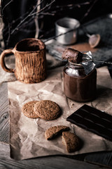 rustic, dark and blurry background. grain. On the table is a jar of chocolate nut paste, a mug and oatmeal cookies.