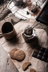 rustic, dark and blurry background. grain. On the table is a jar of chocolate nut paste, a mug and oatmeal cookies.