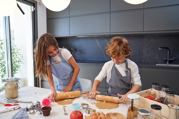 From above of male and female little kids in aprons smiling while rolling brown dough with pin on table at modern kitchen